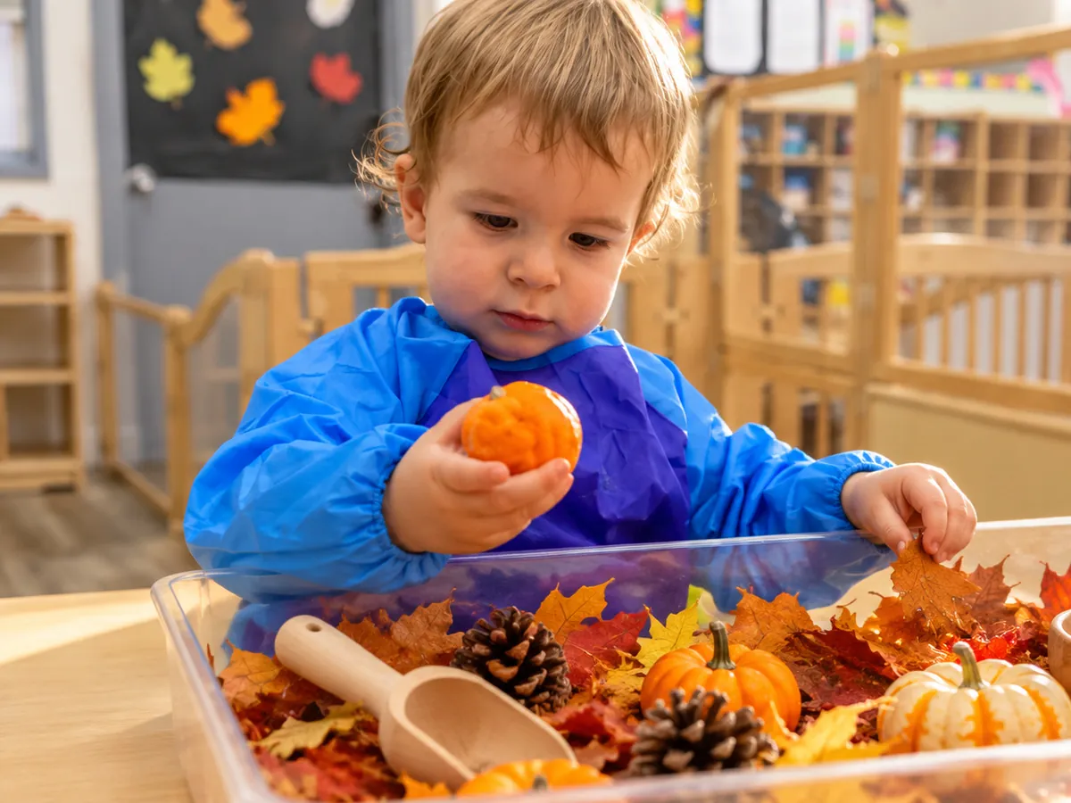 Little hands exploring a fall sensory bin with orange pom-poms, leaves and foam