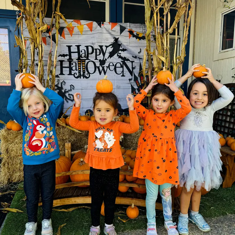Four preschoolers balancing mini pumpkins on their heads in front of the fall photo backdrop