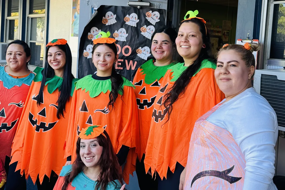 Seven smiling Building Kidz San Jose teachers in matching pumpkin costumes outside a classroom